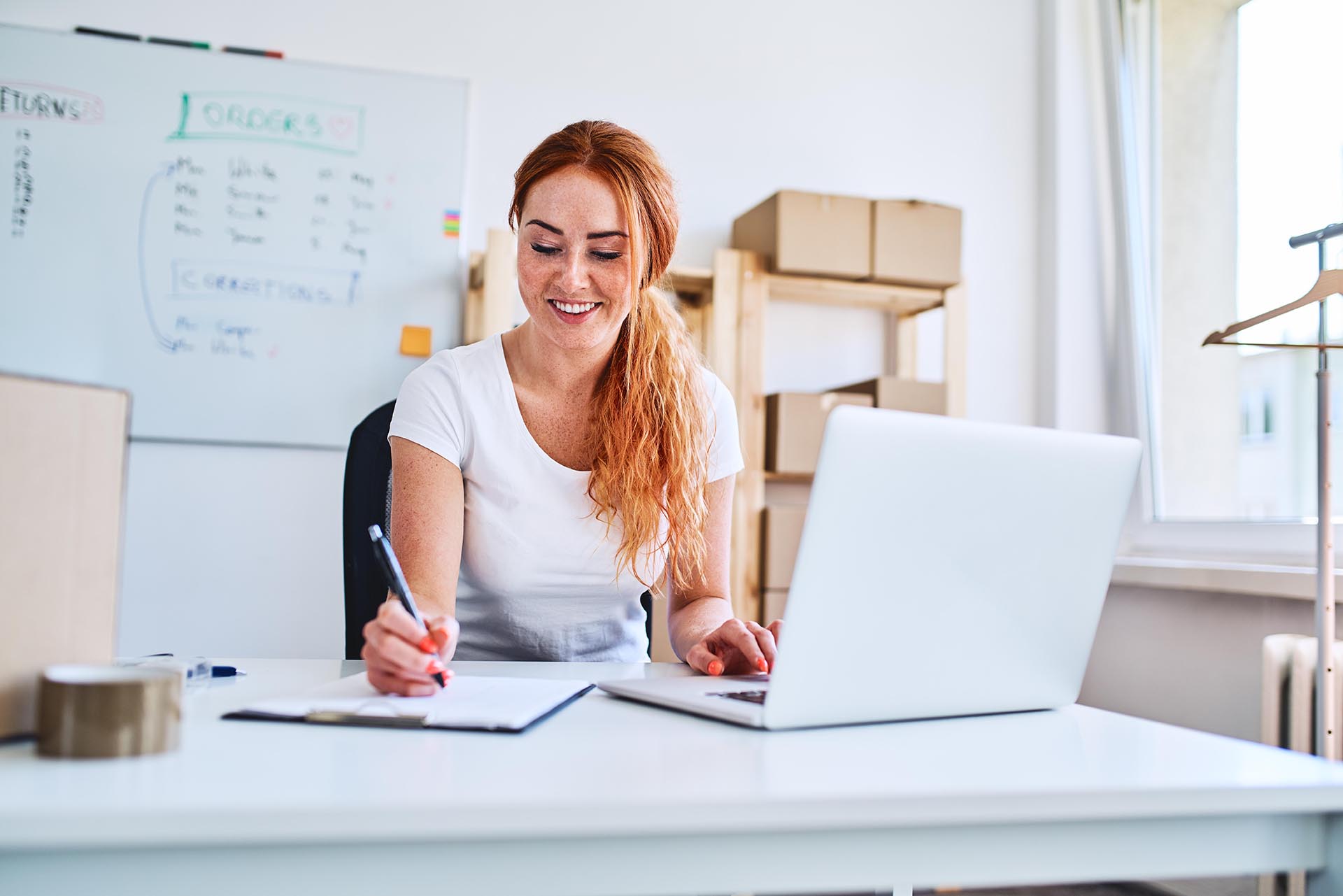 Young female business owner sitting in office writing sales and deliveries on paper sheets while using laptop